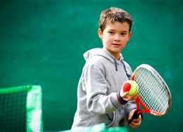A boy wearing a grey hoodie holding a red tennis racket and red and yellow tennis ball