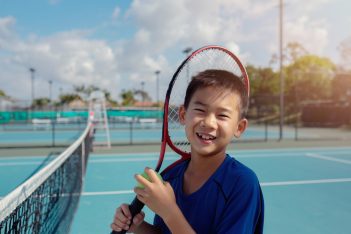 A smiling boy holding a tennis ball and red racket on a tennis court