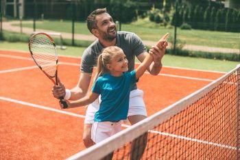 A man helping a girl point at the tennis ball after she hit it