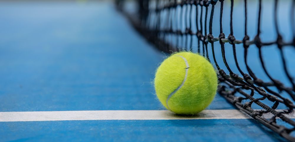 A blue hard tennis court with a black net and tennis ball